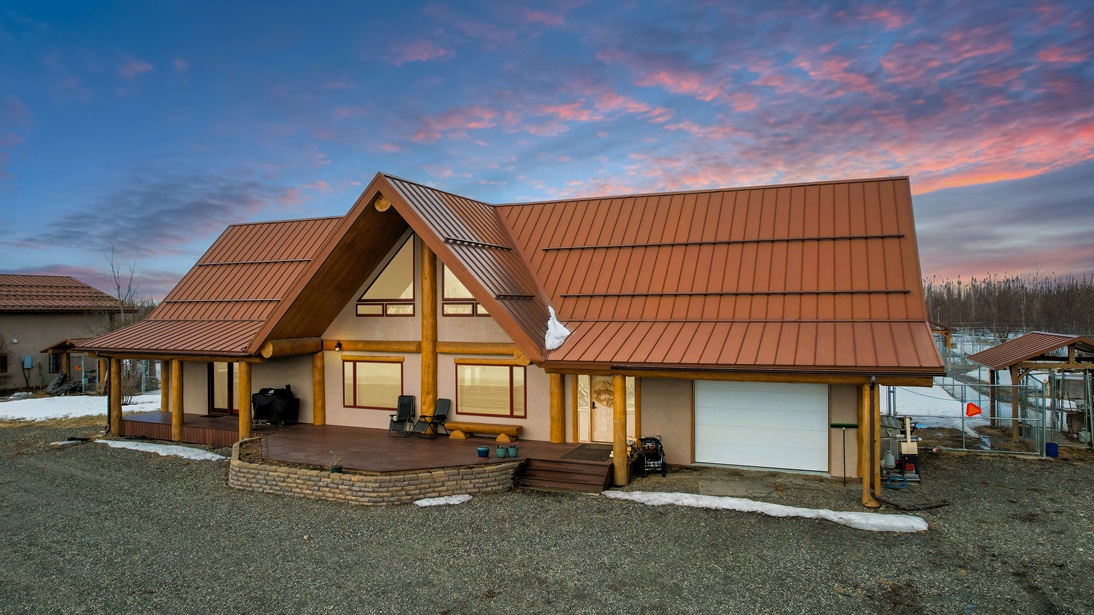 Rustic log cabin-style house with a steep red metal roof, wooden columns, and a raised porch at sunset.