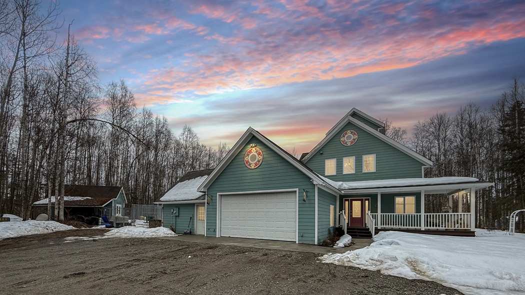 Two-story teal house with a white garage door in a snowy yard at sunset, surrounded by bare trees.