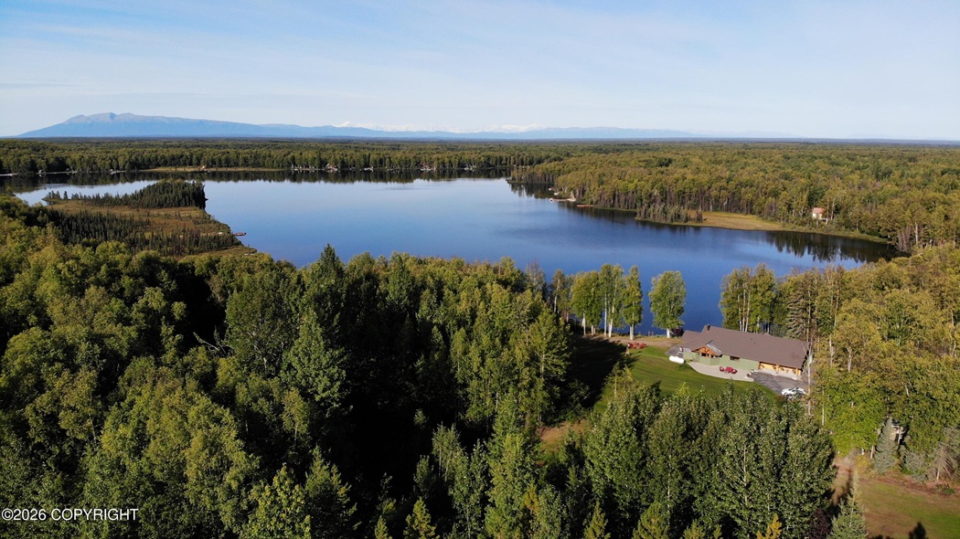 Aerial view of a calm lake surrounded by dense forest, with a lawn and cabin on the shore to the right and distant mountains on the horizon.